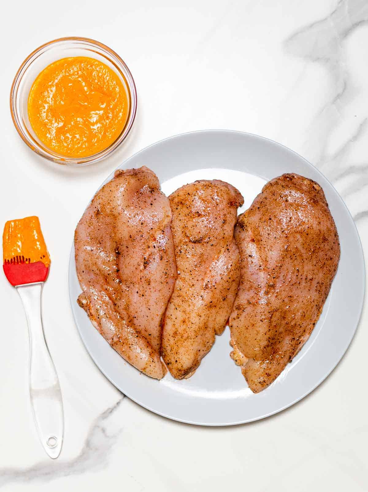 3 chicken breasts rubbed with spices on a grey dish alongside a brush