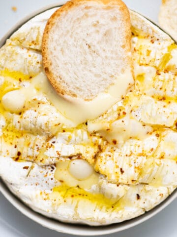 overhead shot of a camembert wheel baked, and drizzled with olive oil on top and a slice of bread