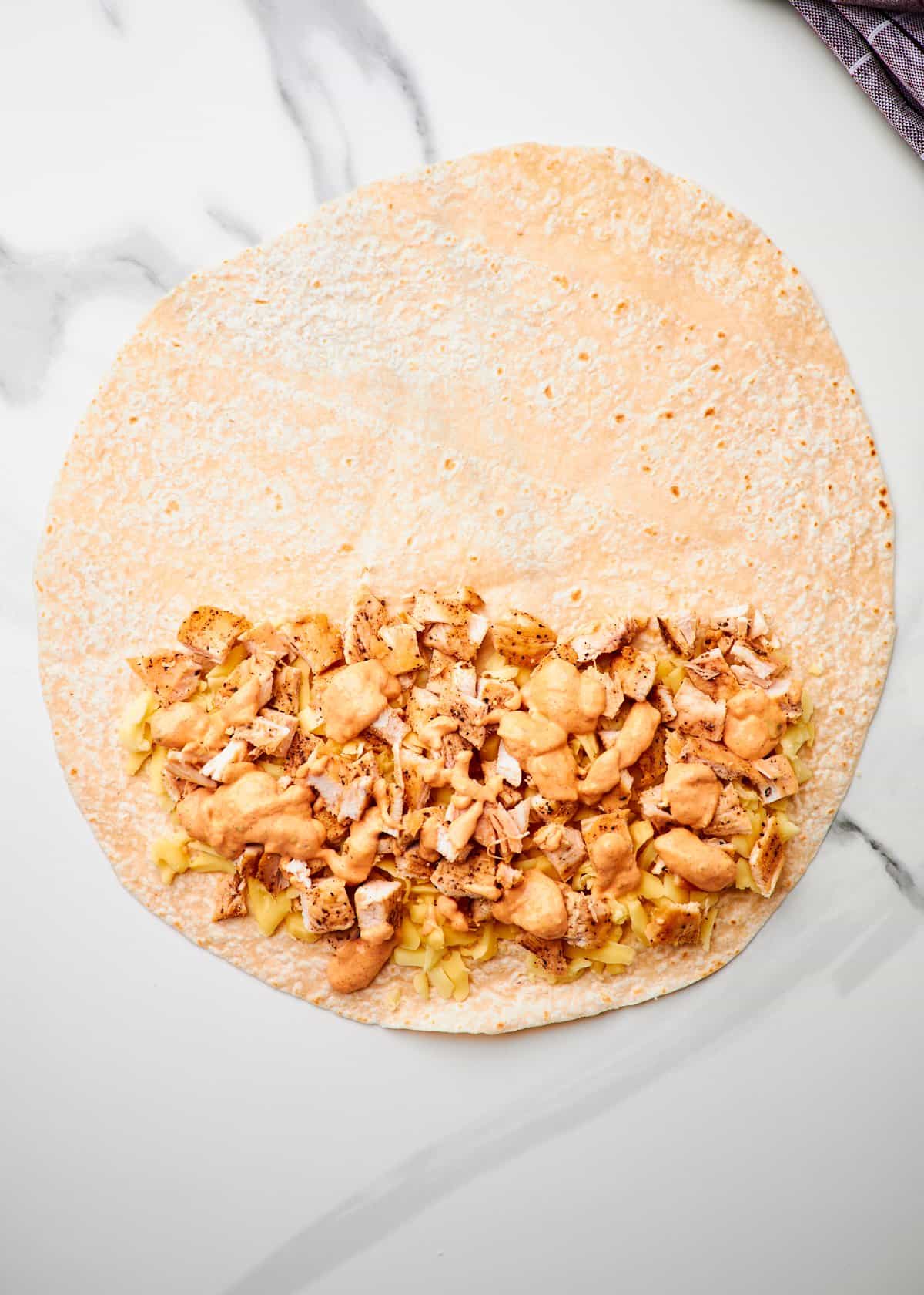 A flour tortilla with shredded cheese, diced grilled chicken, and creamy quesadilla sauce on it.